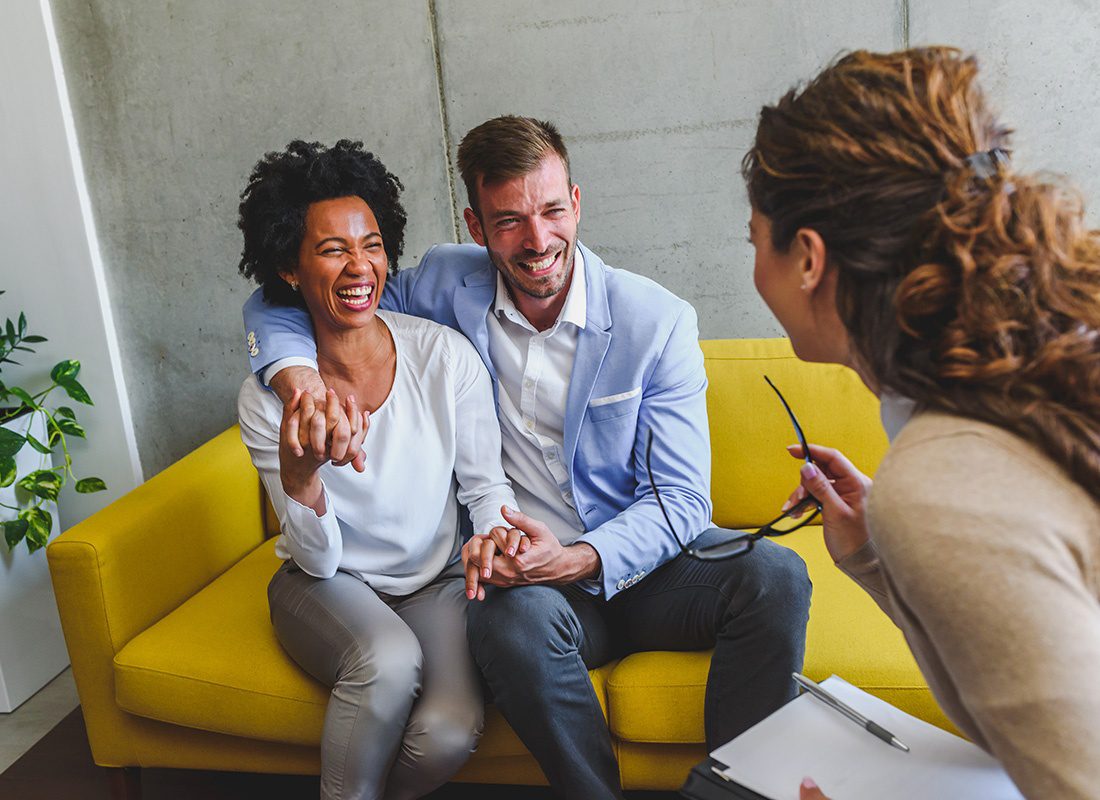 About Our Agency - Cheerful Young Married Couple Sitting on a Yellow Sofa Together in a Modern Office While Talking to Their Female Insurance Agent Holding a Clipboard